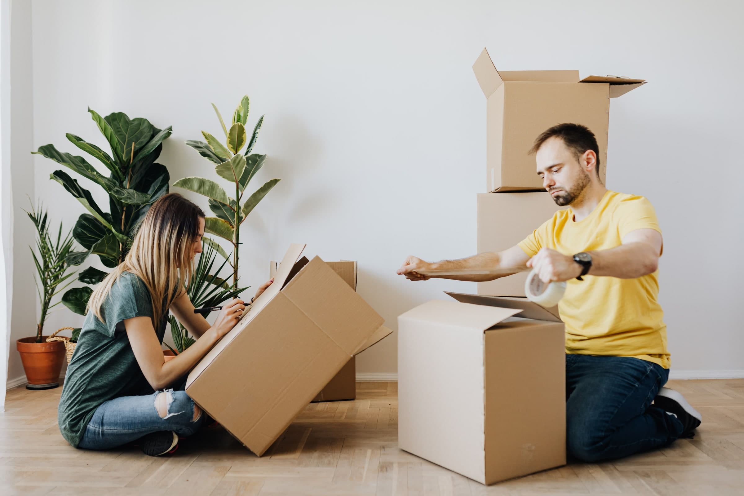 Two people kneeling on a bare floor, labelling and taping cardboard boxes in an apartment being packed up