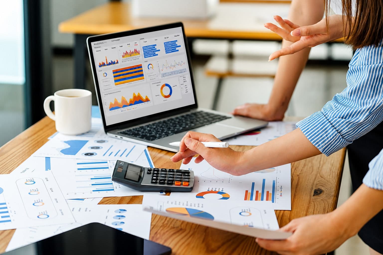 Two colleagues reviewing an analytics dashboard on a laptop, beside printed charts and a calculator